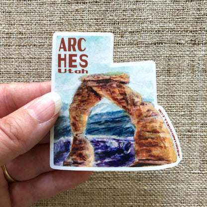 Closeup image of hand holding an Arches National Park, Utah collectible vinyl sticker. Featuring the Delicate Arch in watercolor  with red text that reads Arches Utah. Jhermanrussell.com